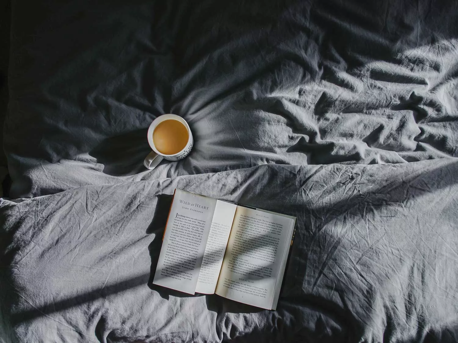 A book and a cup of coffee atop gray sheets.
